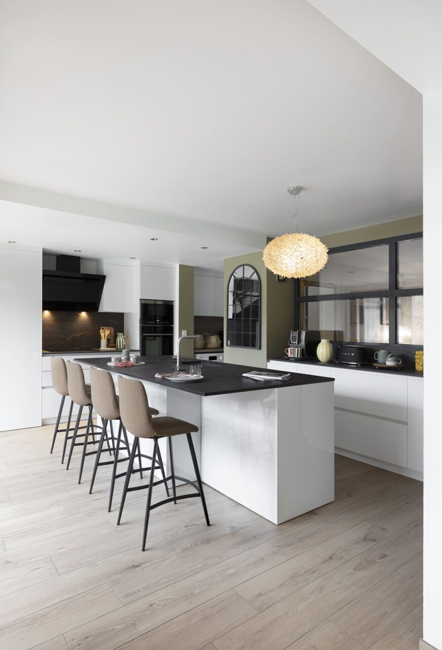 Vue générale de la cuisine rénovée avec grand îlot central, façades blanches et plan de travail noir à Sautron.
